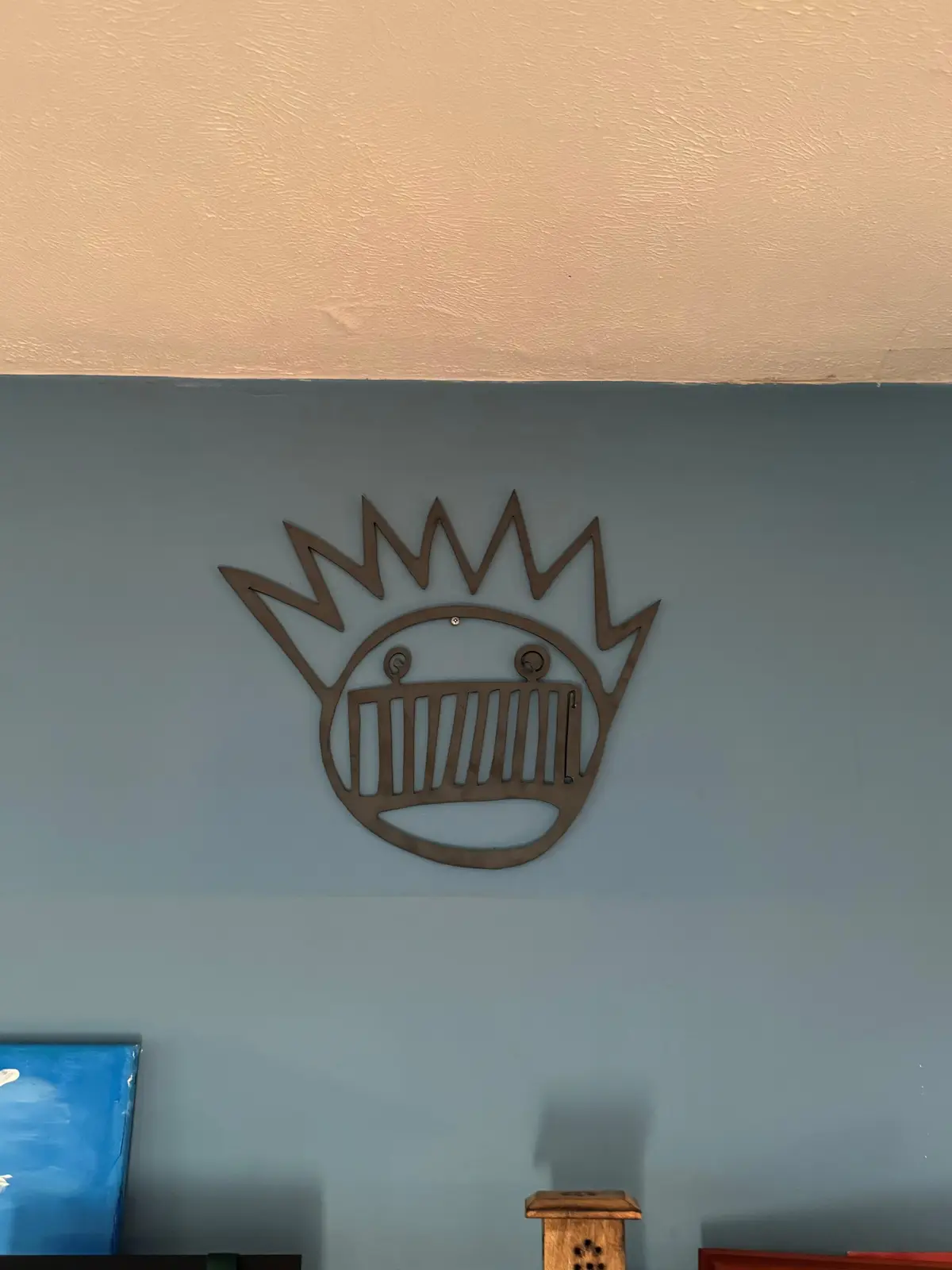 A brown metal wall art piece shaped like a smiling sun with a grill-like mouth is mounted on a two-toned wall, beige above…