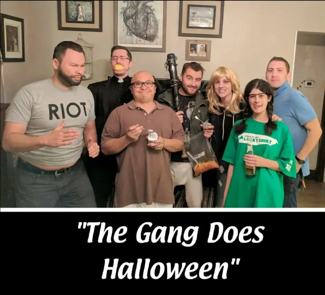 A group of seven people pose indoors for a Halloween photo, with one man in a priest costume holding a lemon, another in a…