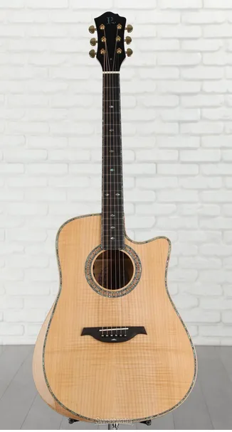 A full-body photo of an acoustic guitar with a cutaway body shape, standing upright against a white brick wall. The guitar…