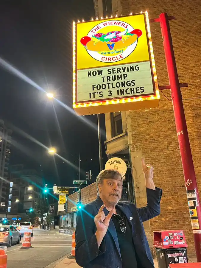 A man stands on a city sidewalk at night, pointing upward toward a brightly lit sign for 'The Wieners Circle' hot dog stand…