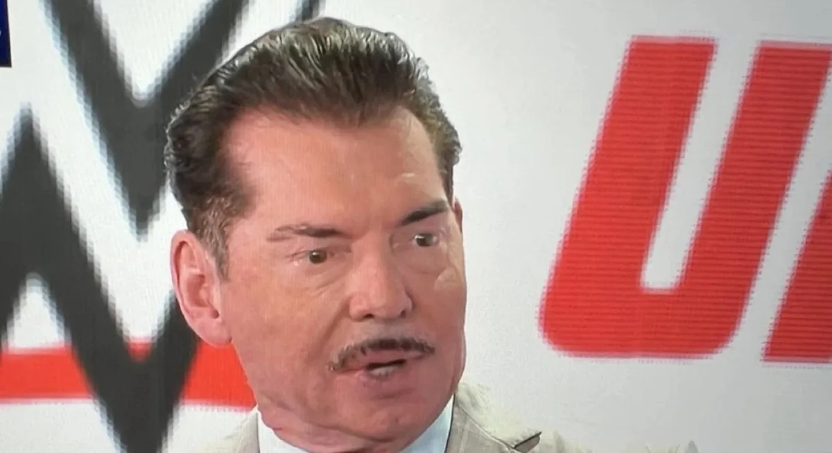 A close-up of a man with dark hair and a mustache, wearing a light-colored suit jacket over a collared shirt, speaking with…