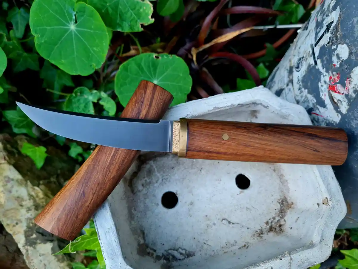 A handmade knife with a wooden handle and brass bolster rests on a weathered concrete block, next to its matching wooden…