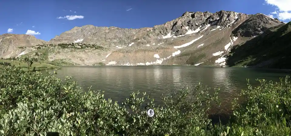 A scenic mountain lake with snow-dusted peaks under a blue sky, framed by green bushes in the foreground. A small circular…