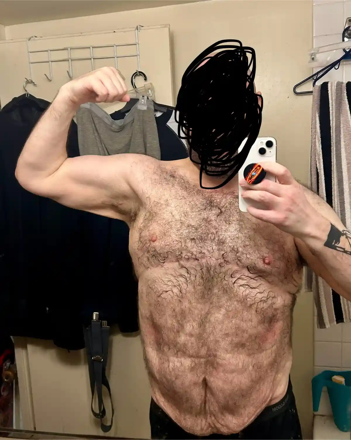 A shirtless man with a hairy chest and defined abs takes a mirror selfie in a bathroom, flexing his bicep while holding a…