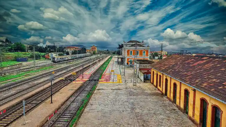 An empty train station with multiple tracks, platforms, and old buildings under a dramatic, cloudy sky. A train is visible…