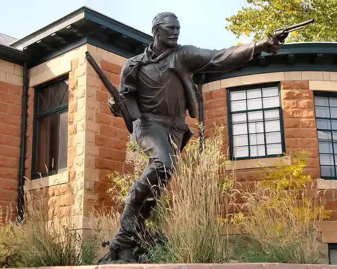 A bronze statue of a man in rugged attire, holding a rifle in one hand and pointing a pistol with the other, stands outdoors…