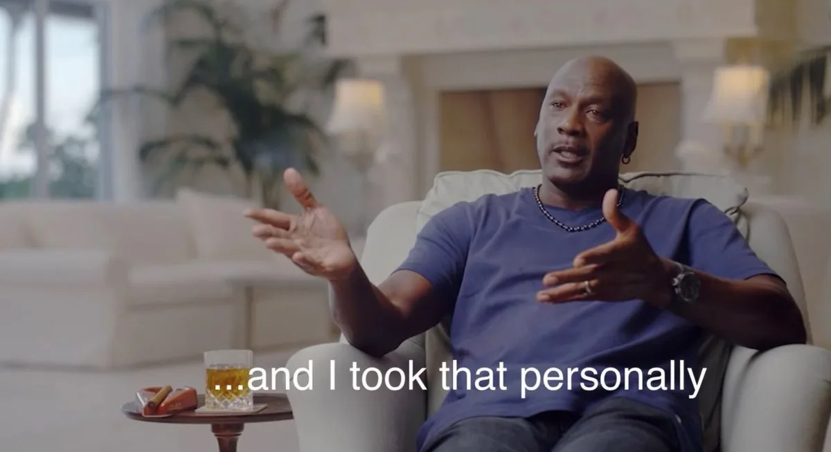 Michael Jordan sits in a white armchair in a luxurious living room, gesturing with both hands while speaking. A glass of…