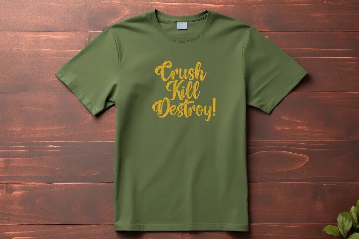 An olive green t-shirt with yellow cursive text reading 'Crush Kill Destroy!' is laid flat on a dark wooden surface. The…