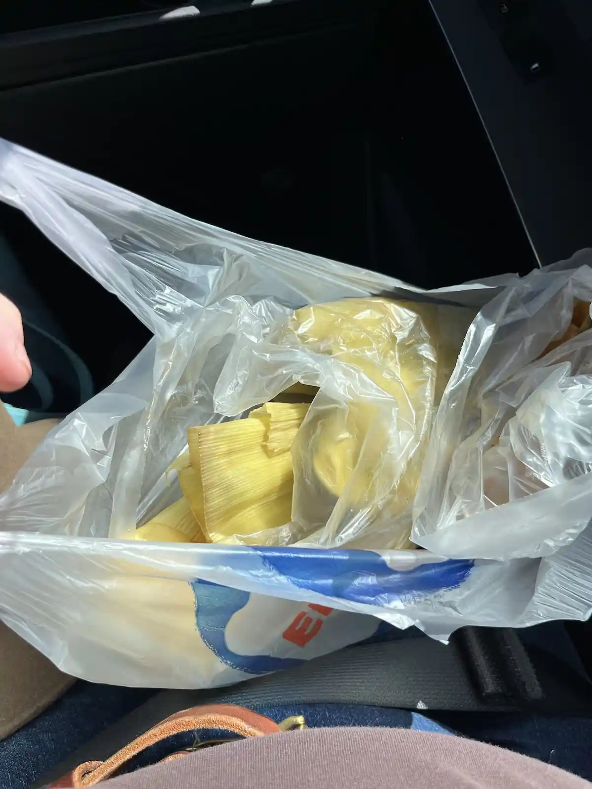 A person holds open a clear plastic grocery bag inside a car, revealing yellow corn husks inside. The bag has a blue and red…