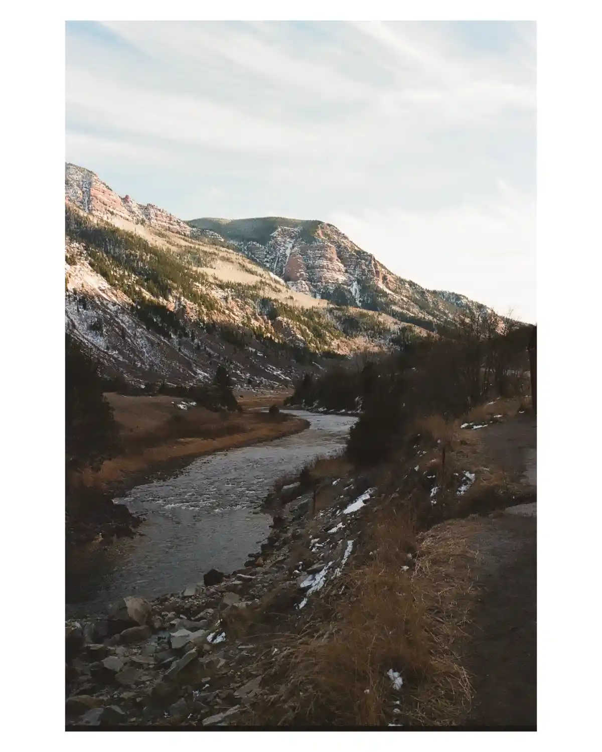 A scenic photo of a river flowing through a mountain valley with snow-dusted slopes and evergreen trees under a partly…