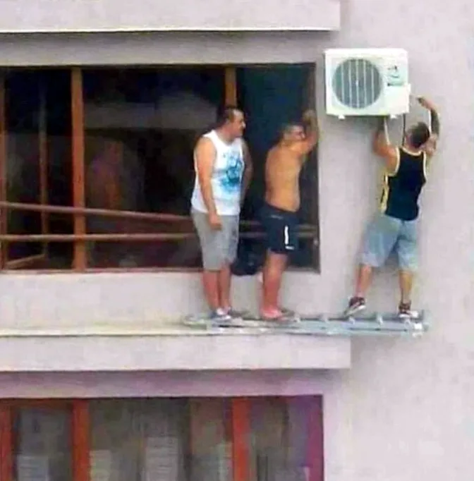 Three men stand on a narrow metal platform attached to the side of a building, attempting to install an outdoor air…