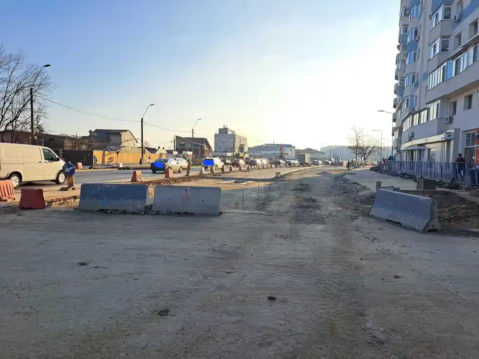 A street under construction with dirt, concrete barriers, and orange traffic cones. Cars drive on the remaining paved road…