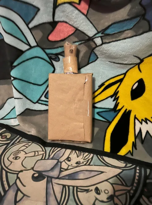 A handmade, rectangular brown paper package with a taped top sits on a colorful blanket featuring various Pokémon…