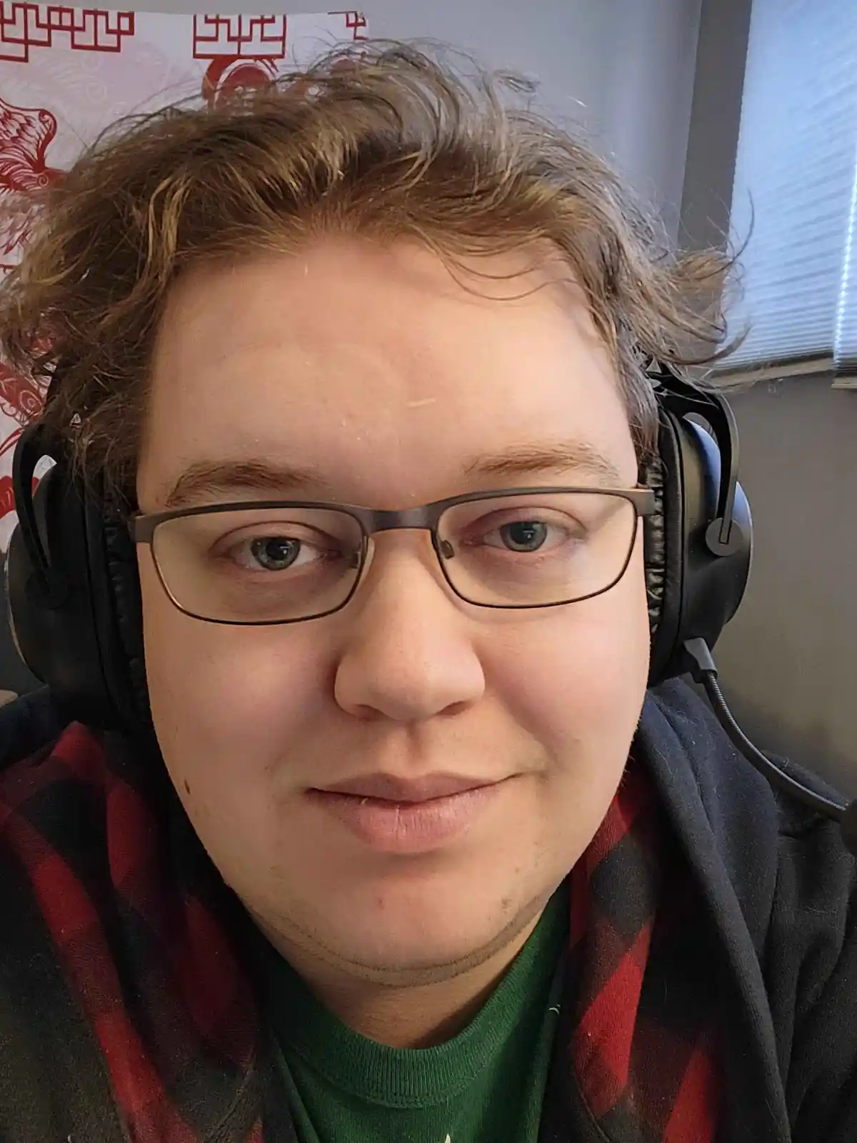 A person with curly brown hair, wearing glasses and black over-ear headphones, looks directly at the camera with a subtle…