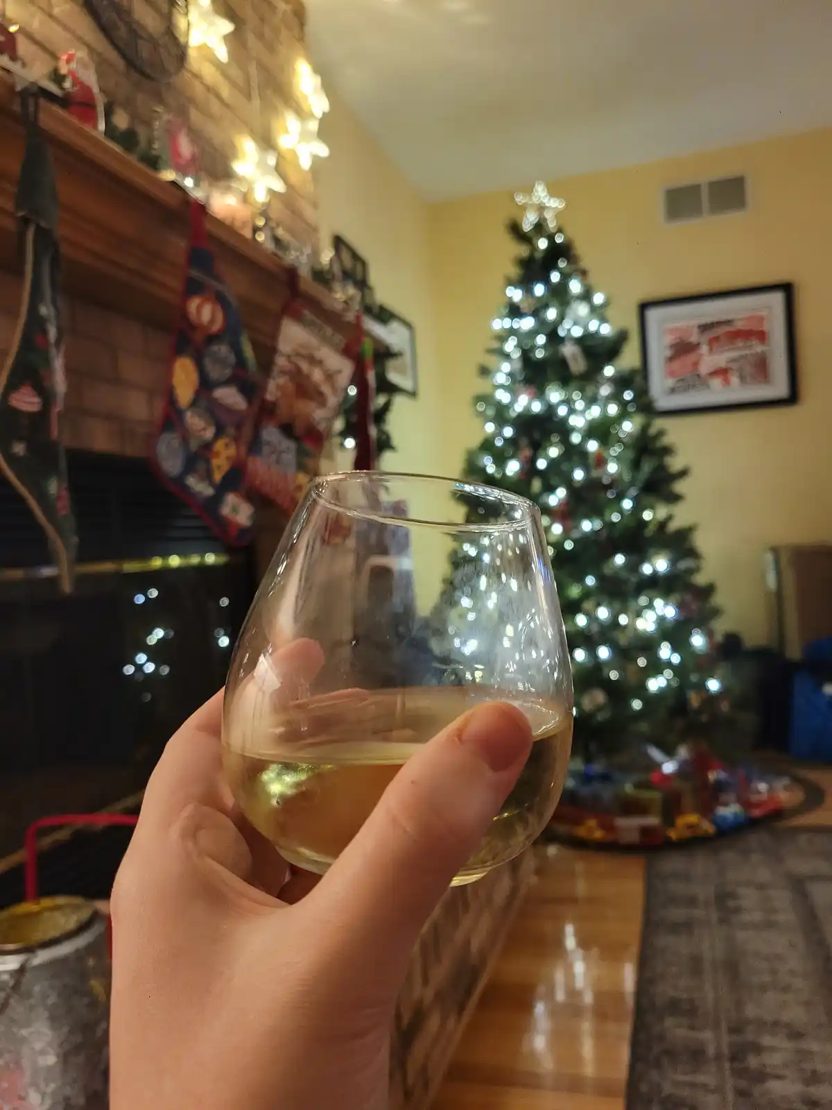 A hand holds a nearly empty glass of white wine in front of a decorated christmas tree and fireplace, with the overlaid text…