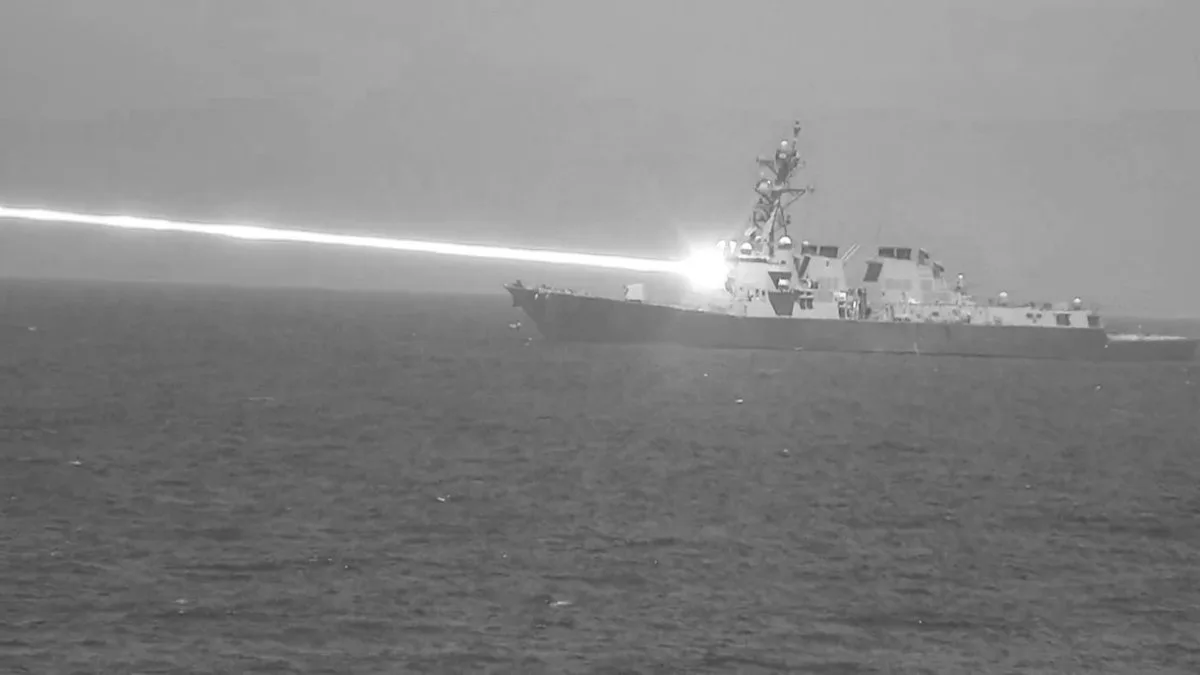 A black-and-white image shows a naval warship on the ocean, emitting a bright, long, horizontal beam of light from its…