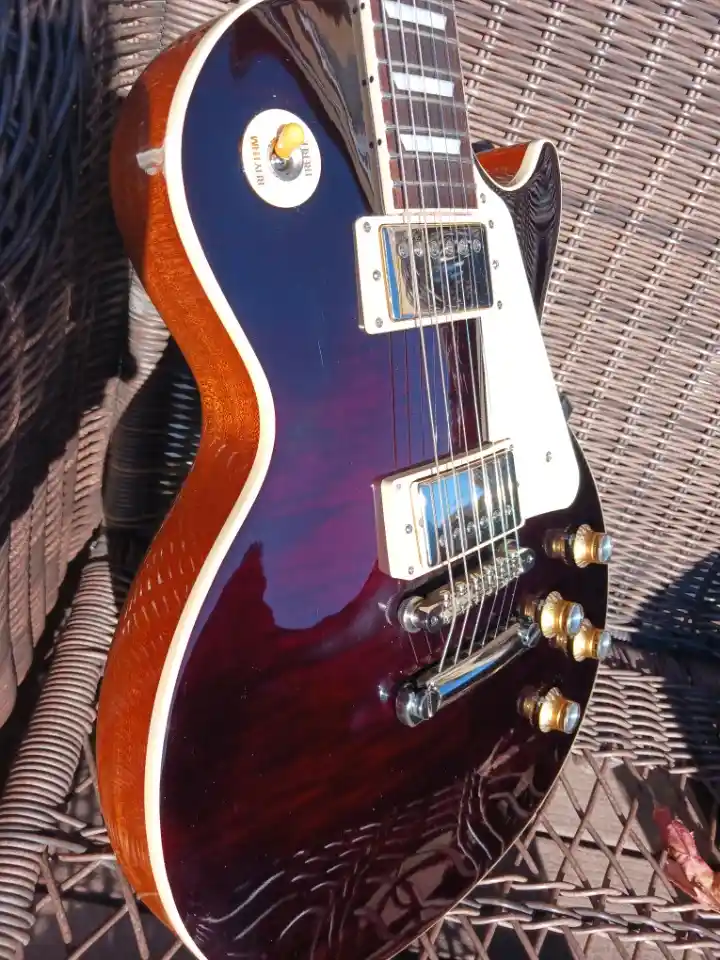 A close-up photo of a dark purple electric guitar with a glossy finish, resting on a brown wicker chair. The guitar has a…