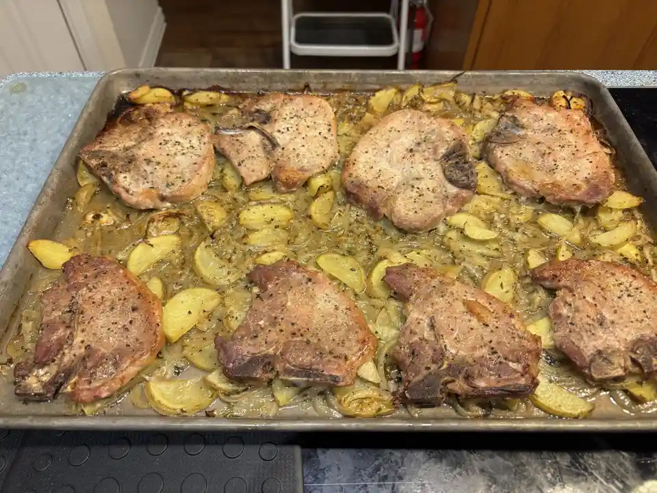A baking sheet holds eight cooked pork chops and potato wedges, with the top row appearing lightly browned and the bottom…