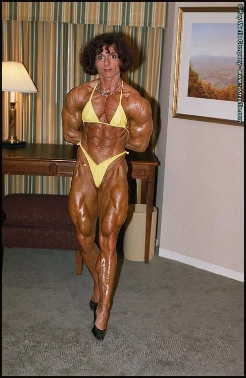 A woman with extremely muscular, oiled physique poses in a yellow bikini and high heels in a hotel room. She stands in front…