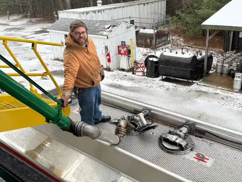 A man in an orange hoodie and glasses stands on the metal deck of a fuel tanker truck, holding a green fueling hose. Behind…