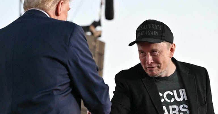elon musk wearing a black 'make america great again' cap and a partially visible t-shirt, looking toward donald trump who is…