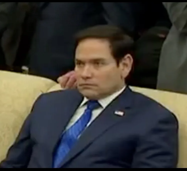 a man in a dark blue suit and blue tie sits on a beige couch with a stern, unimpressed facial expression. His right hand is…