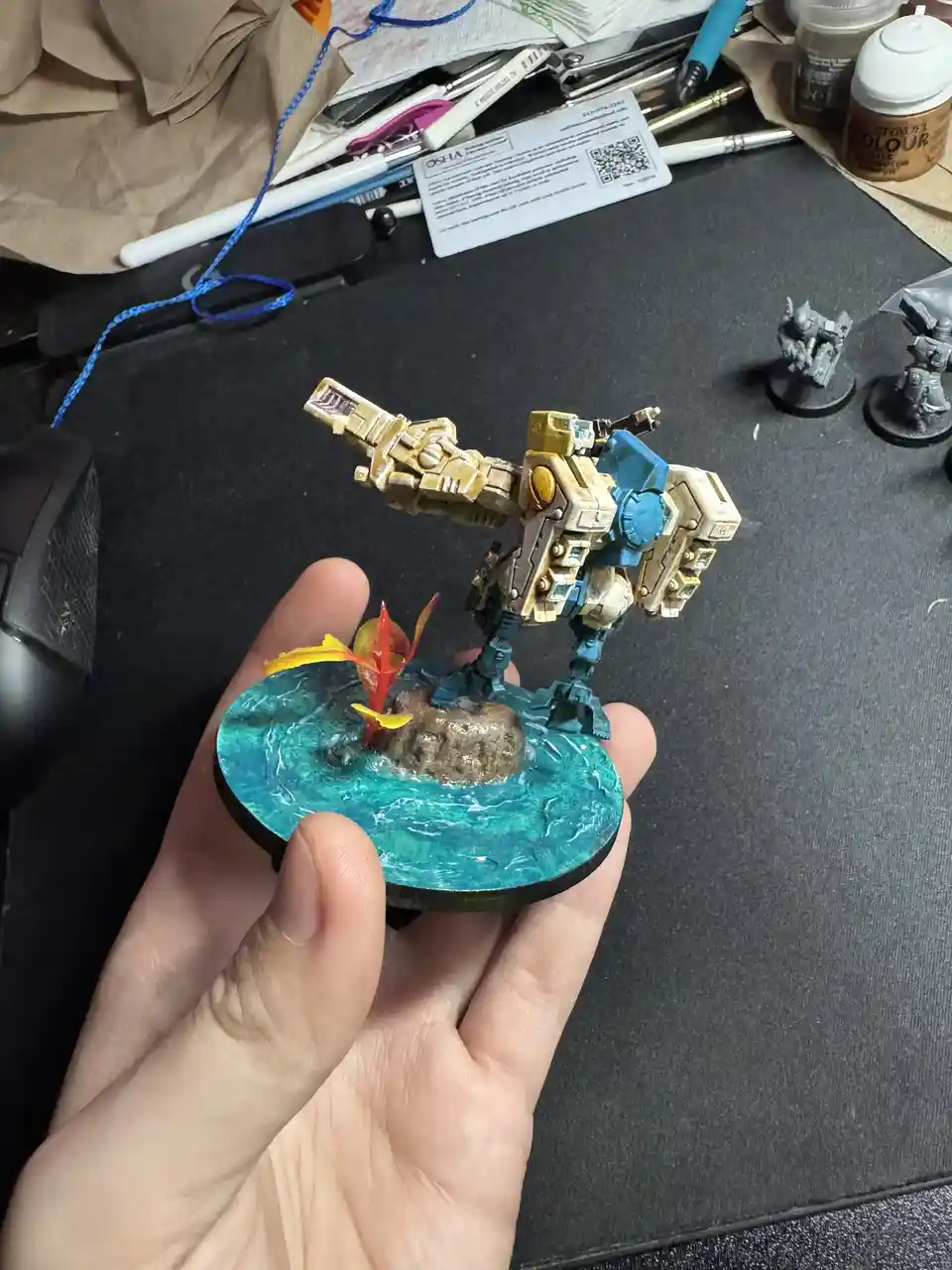 A hand holds a painted miniature of a large, armored robot standing on a circular base that has been meticulously painted to…