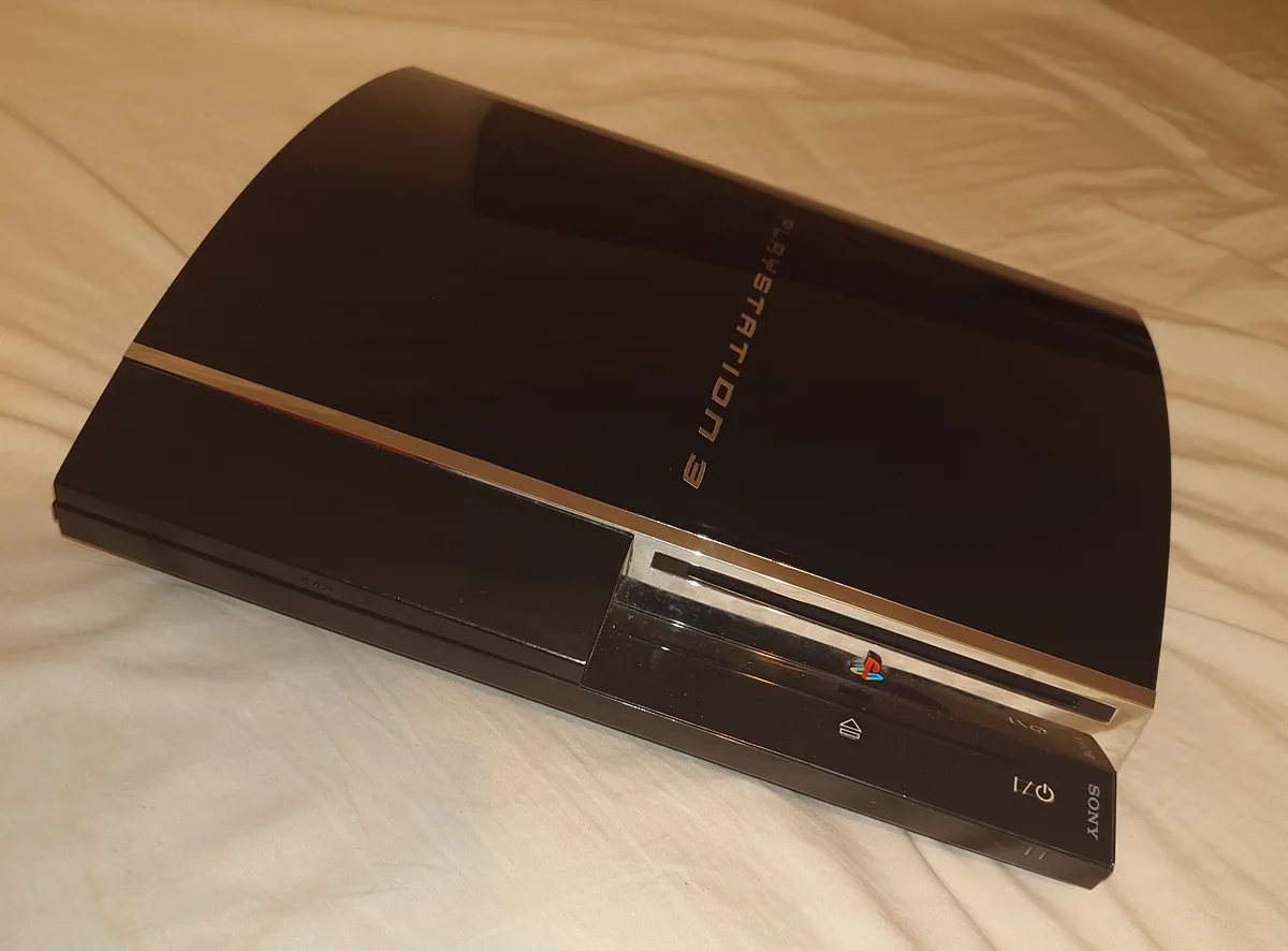 A black PlayStation 3 console with a gold stripe lies on a white bedsheet, its 'PLAYSTATION 3' branding visible. The…