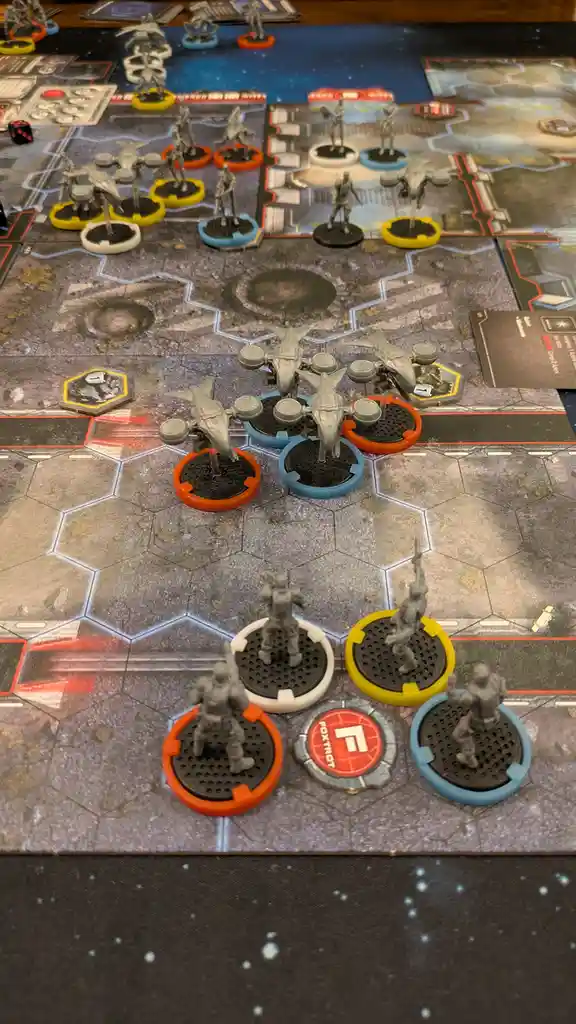 A tabletop wargame setup shows numerous grey plastic miniatures on colored circular bases arranged on a hexagonal grid map…
