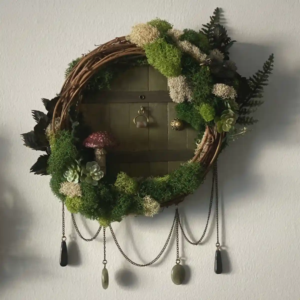 A circular wreath made of woven branches, green and beige moss, ferns, and succulents frames a miniature wooden door with a…