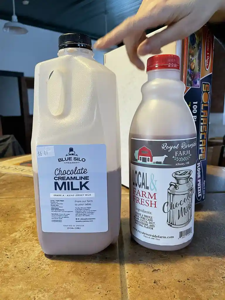 A hand points at a large jug of Blue Silo Creamery chocolate creamline milk, priced at $6.49, standing next to a smaller…