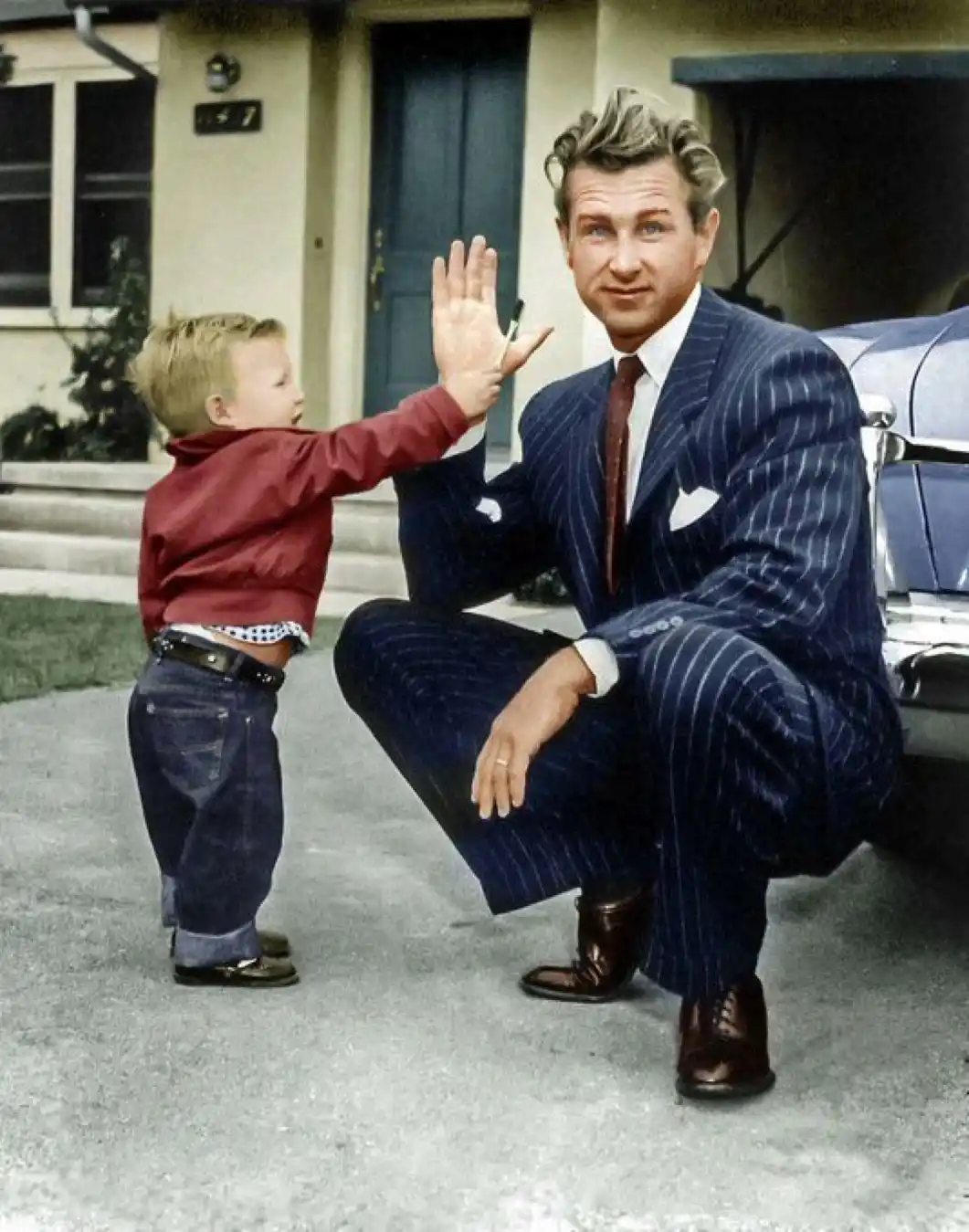 A vintage photo shows a young boy in a red shirt and jeans high-fiving a man in a pinstripe suit who is crouching down to…