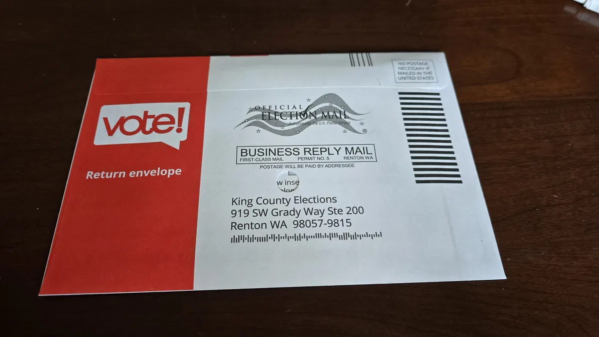 A red and white official election mail return envelope from King County Elections in Renton, WA, sits on a dark wooden…