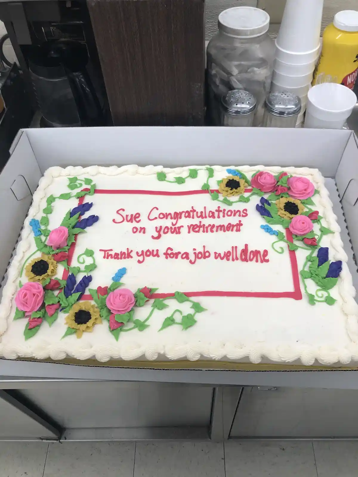 A rectangular white frosted cake in a cardboard box, decorated with pink roses, yellow sunflowers, and green vines, sits on…