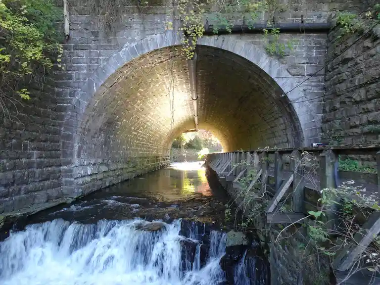 A photo of a stone arch tunnel with a stream flowing through it, leading to bright sunlight at the end. A wooden walkway…