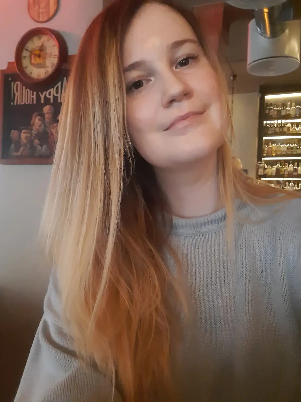 A young woman with long blonde hair and a light grey sweater smiles subtly at the camera in what appears to be a bar, with a…
