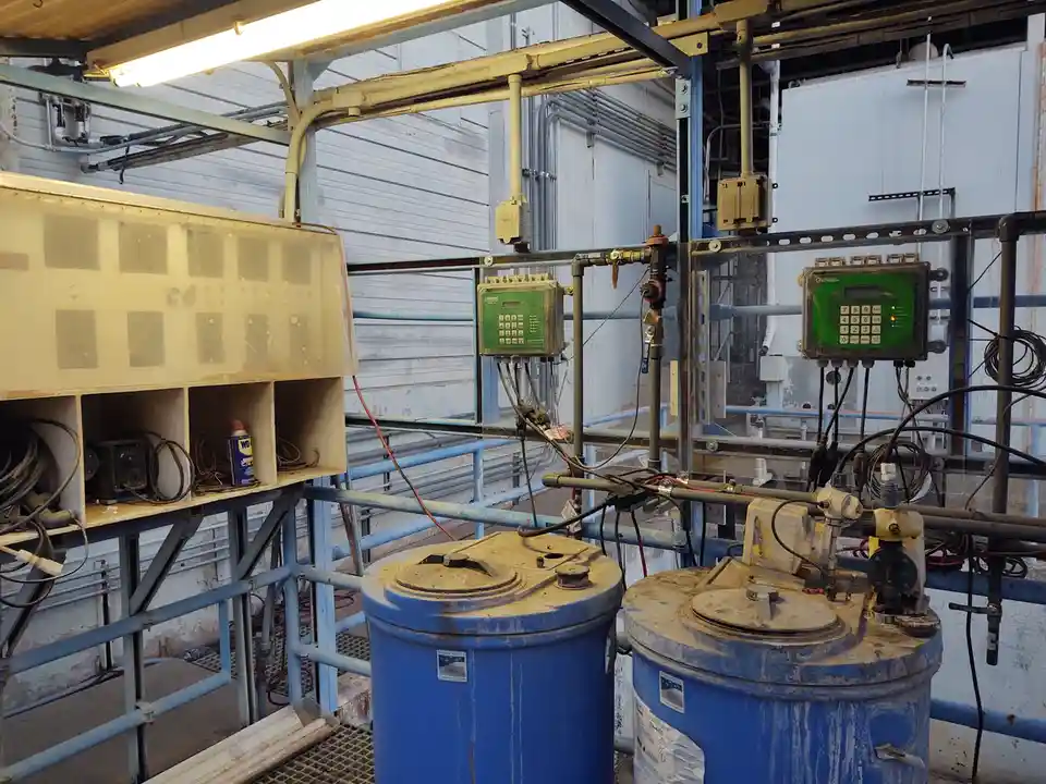 An industrial setting with two large blue barrels, pipes, and control panels, one of which has a green keypad. A can of…