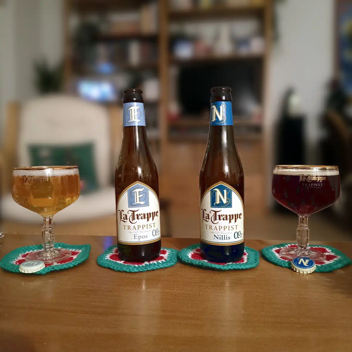 Two La Trappe Trappist beer bottles, labeled 'Epos' and 'Nillis', each with 0.9% alcohol, sit on a wooden table with…
