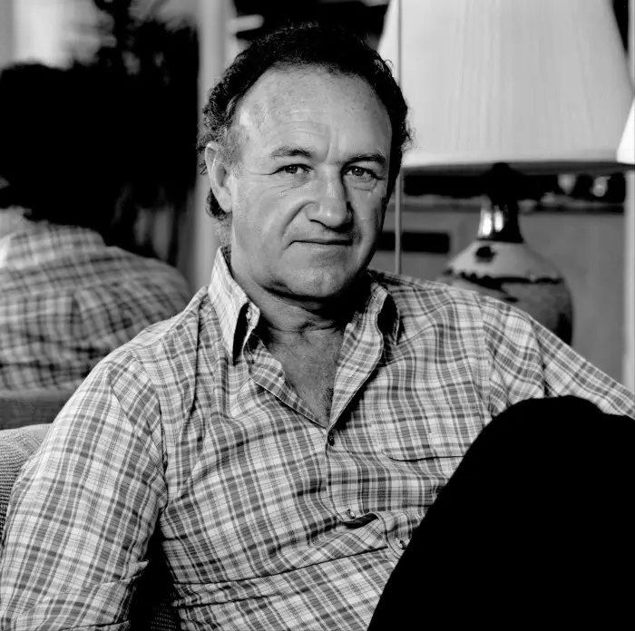 A black-and-white photo of actor Gene Hackman sitting in a chair, wearing a plaid shirt, with a subtle, knowing smirk. The…