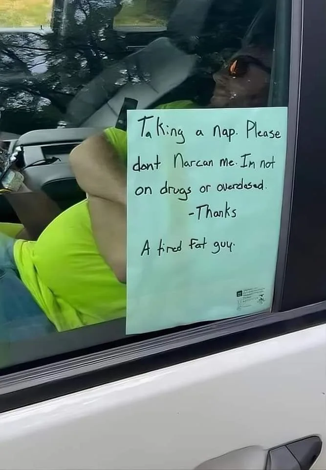 A person in a bright green shirt is seen sleeping inside a white car, with a handwritten note taped to the window. The note…