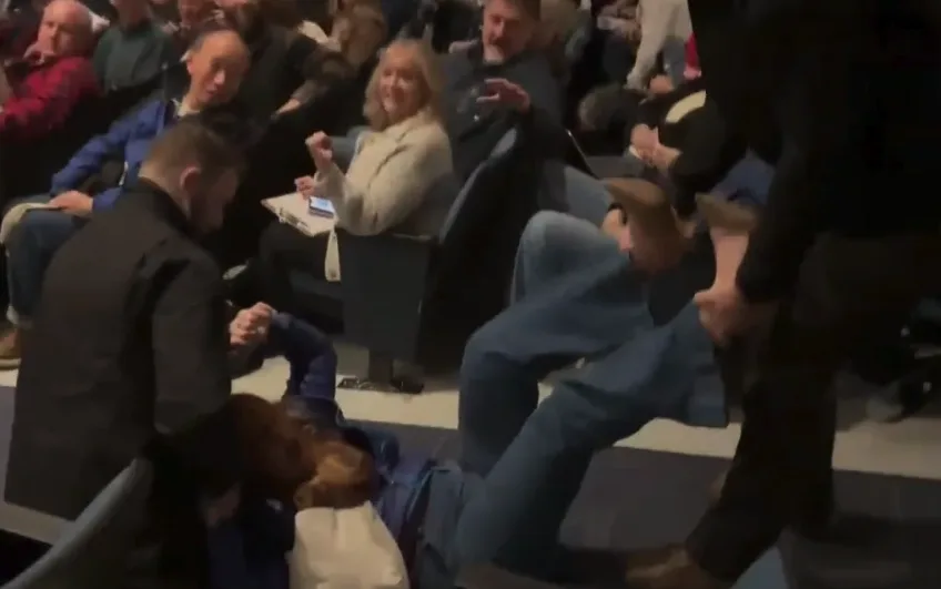 A person is lying on the floor in front of an audience, while a man in a black jacket kneels beside them. Other audience…