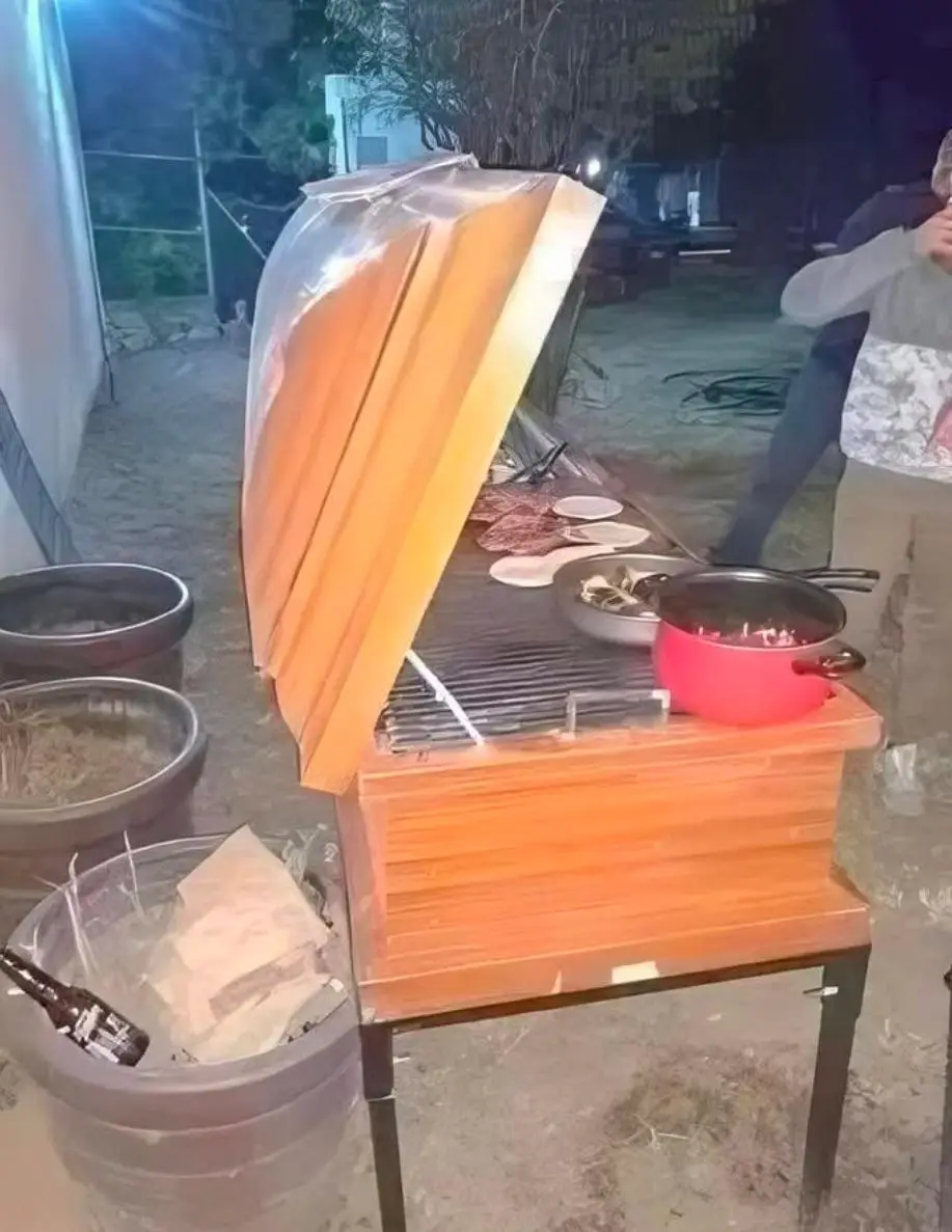 a wooden outdoor grill at night with its lid propped open and covered by a clear plastic bag, a red pot sits on the grill…
