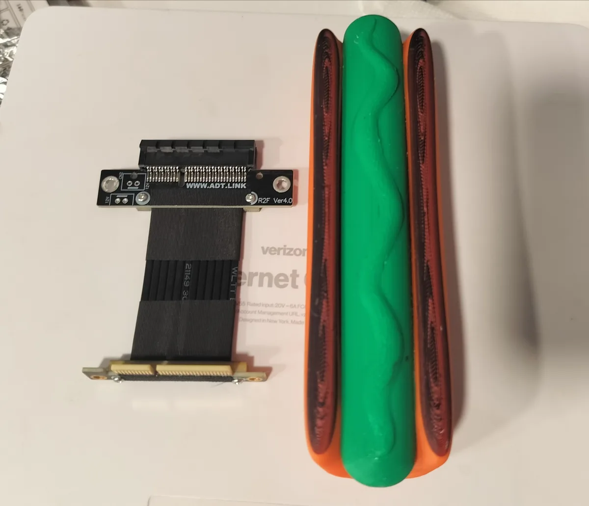 An image shows a computer PCIe riser cable adapter next to a 3D-printed object shaped like a hot dog with two buns, one…