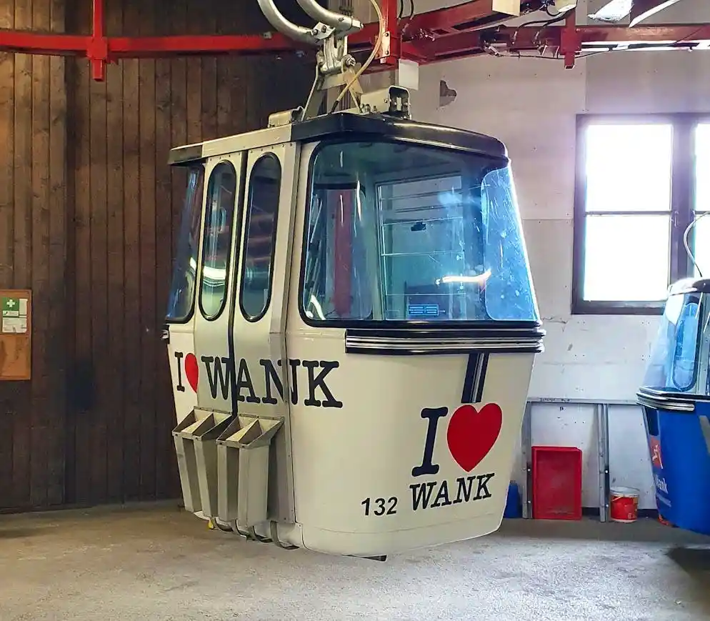 A white cable car cabin, numbered 132, is parked indoors with the phrase 'I ❤️ WANK' painted in large black letters and a…