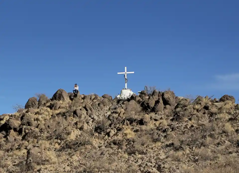 A person sits atop a rocky, arid hillside under a clear blue sky, with a large white cross mounted on a stone base visible…