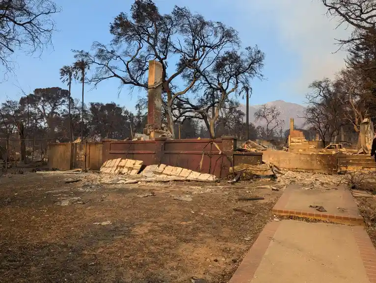 A desolate landscape shows the charred remains of a house after a wildfire, with a lone chimney standing amidst rubble…