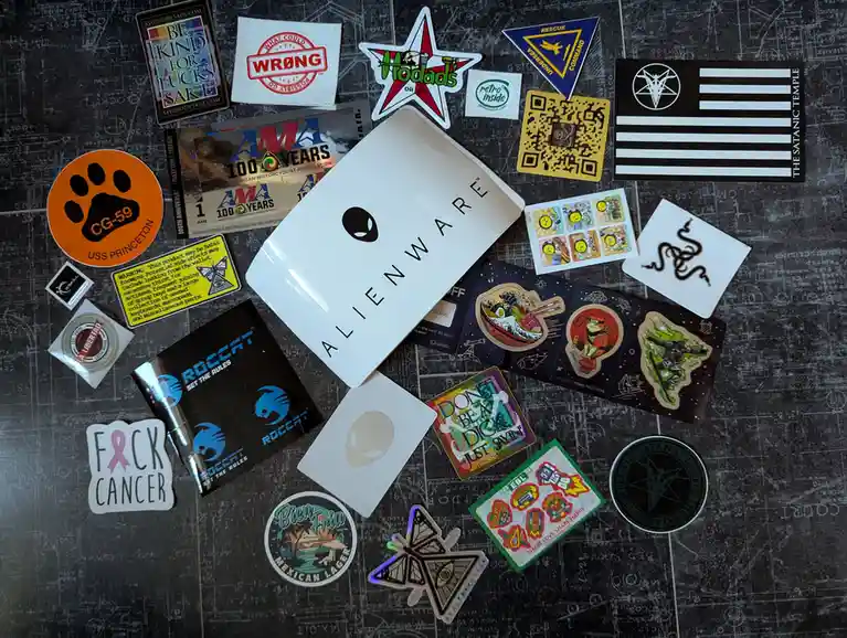 A flat lay of numerous stickers and patches scattered on a dark surface covered in faint white engineering-style grid lines…