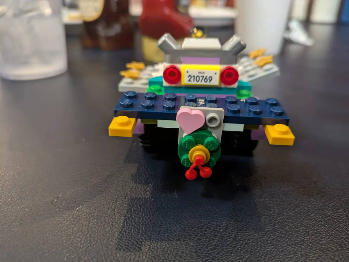 A small, colorful Lego car built from various bricks sits on a dark textured surface. The car has a blue base, green front…