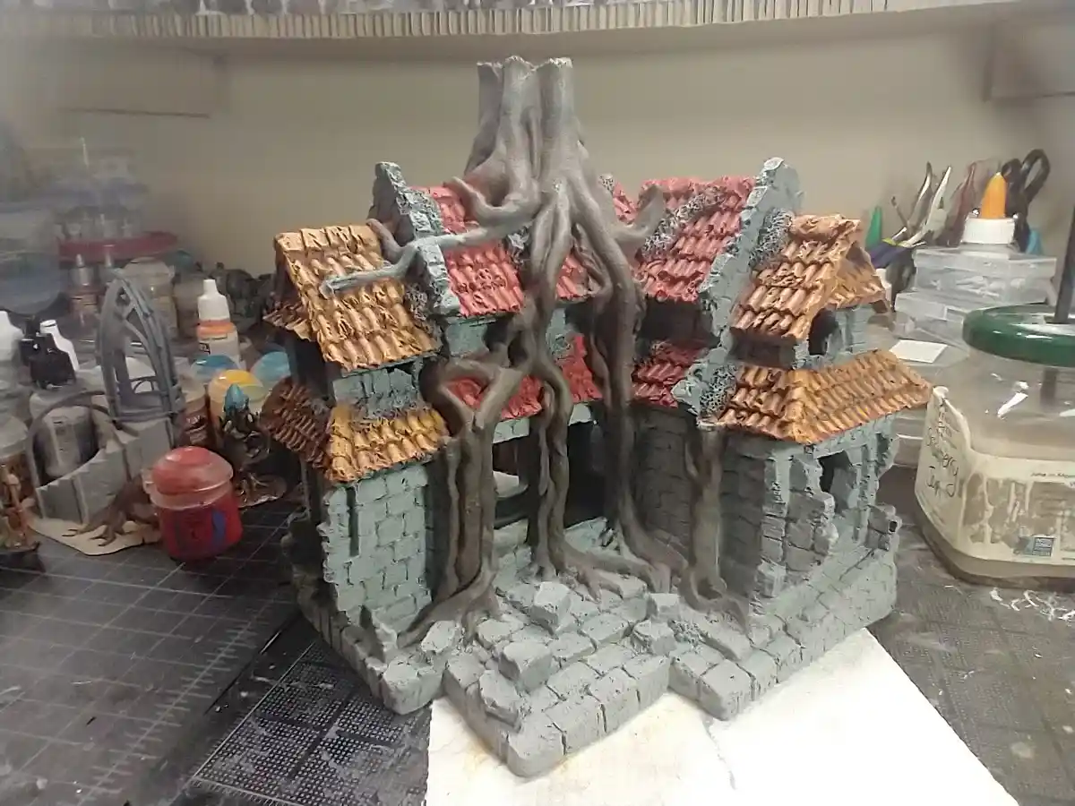 A miniature diorama of a fantasy-style village with crumbling stone walls and red-tiled roofs, overtaken by large, gnarled…