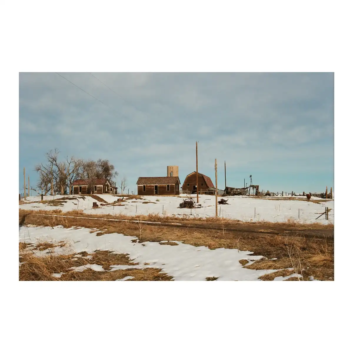 A snowy, rural landscape features dilapidated wooden farm buildings, including a barn and house, surrounded by bare trees…